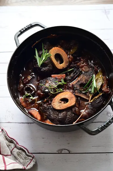 Yanında taze patates, kök sebze ve taze otlarla birlikte kırmızı şarap soslu kemikli haşlanmış sığır eti servis edilir. Beyaz bir tabakta parlak bir arkaplanda sunulan bu çanak, klasik konforlu yiyecekleri inceliyor.