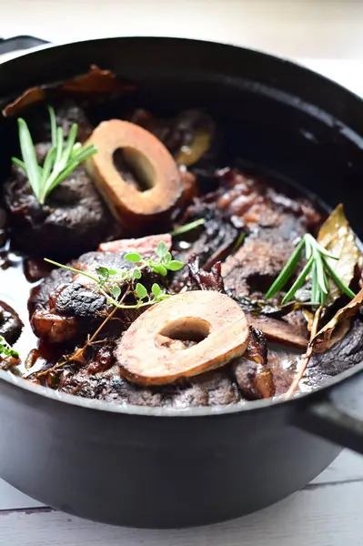 Yanında taze patates, kök sebze ve taze otlarla birlikte kırmızı şarap soslu kemikli haşlanmış sığır eti servis edilir. Beyaz bir tabakta parlak bir arkaplanda sunulan bu çanak, klasik konforlu yiyecekleri inceliyor.