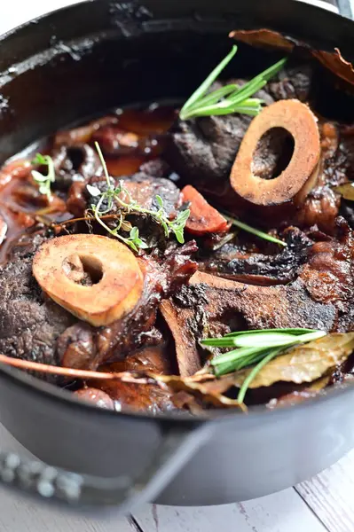 Yanında taze patates, kök sebze ve taze otlarla birlikte kırmızı şarap soslu kemikli haşlanmış sığır eti servis edilir. Beyaz bir tabakta parlak bir arkaplanda sunulan bu çanak, klasik konforlu yiyecekleri inceliyor.