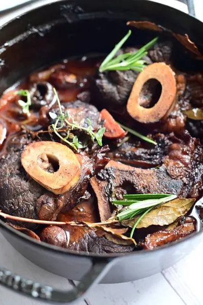 Yanında taze patates, kök sebze ve taze otlarla birlikte kırmızı şarap soslu kemikli haşlanmış sığır eti servis edilir. Beyaz bir tabakta parlak bir arkaplanda sunulan bu çanak, klasik konforlu yiyecekleri inceliyor.