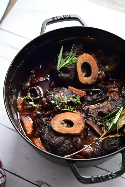 Yanında taze patates, kök sebze ve taze otlarla birlikte kırmızı şarap soslu kemikli haşlanmış sığır eti servis edilir. Beyaz bir tabakta parlak bir arkaplanda sunulan bu çanak, klasik konforlu yiyecekleri inceliyor.