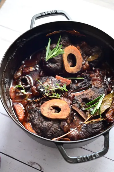 Yanında taze patates, kök sebze ve taze otlarla birlikte kırmızı şarap soslu kemikli haşlanmış sığır eti servis edilir. Beyaz bir tabakta parlak bir arkaplanda sunulan bu çanak, klasik konforlu yiyecekleri inceliyor.