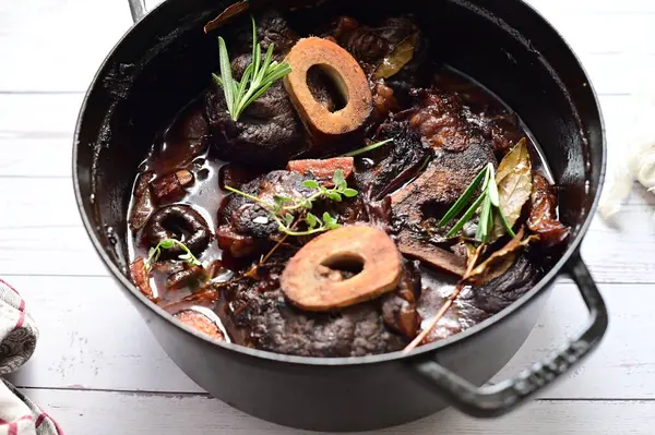 Yanında taze patates, kök sebze ve taze otlarla birlikte kırmızı şarap soslu kemikli haşlanmış sığır eti servis edilir. Beyaz bir tabakta parlak bir arkaplanda sunulan bu çanak, klasik konforlu yiyecekleri inceliyor.