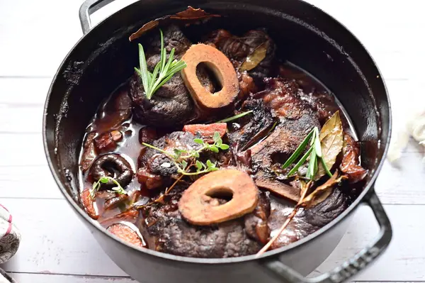 Yanında taze patates, kök sebze ve taze otlarla birlikte kırmızı şarap soslu kemikli haşlanmış sığır eti servis edilir. Beyaz bir tabakta parlak bir arkaplanda sunulan bu çanak, klasik konforlu yiyecekleri inceliyor.