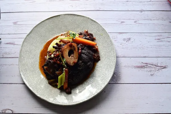 Beyaz bir tabakta renkli sebzelerle servis edilen kemikli kızarmış sığır eti. Tabakta yumuşak, fırında kızartılmış et ve mevsimlik sebzeler yer alıyor ve temiz ve minimalist bir şekilde sunuluyor.