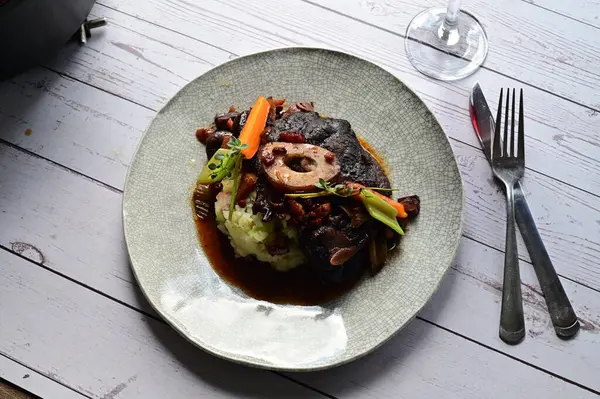 Beyaz bir tabakta renkli sebzelerle servis edilen kemikli kızarmış sığır eti. Tabakta yumuşak, fırında kızartılmış et ve mevsimlik sebzeler yer alıyor ve temiz ve minimalist bir şekilde sunuluyor.