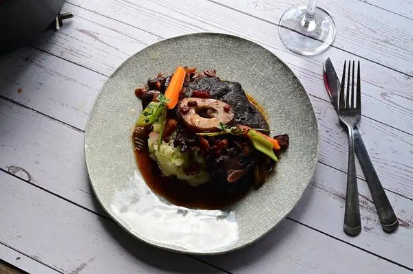 Beyaz bir tabakta renkli sebzelerle servis edilen kemikli kızarmış sığır eti. Tabakta yumuşak, fırında kızartılmış et ve mevsimlik sebzeler yer alıyor ve temiz ve minimalist bir şekilde sunuluyor.