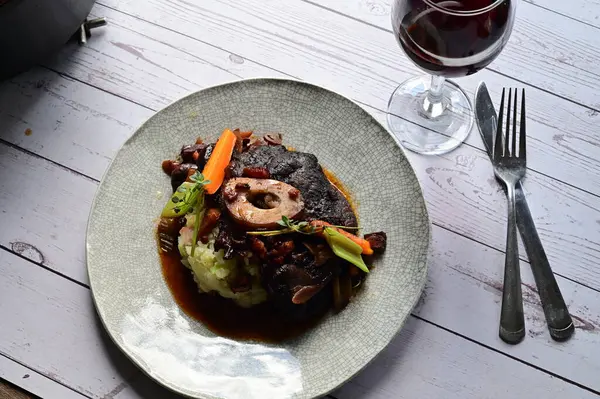 Beyaz bir tabakta renkli sebzelerle servis edilen kemikli kızarmış sığır eti. Tabakta yumuşak, fırında kızartılmış et ve mevsimlik sebzeler yer alıyor ve temiz ve minimalist bir şekilde sunuluyor.