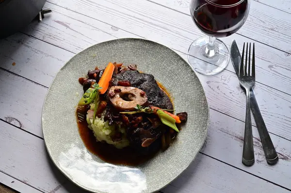 Sebzelerle servis edilen ve taze otlarla süslenen sığır eti osso buco 'sunun yakın çekimi. Kemik iliğiyle pişirilmiş yumuşak et, bu geleneksel İtalyan yemeğinin dokusunu ve derinliğini vurguluyor. 