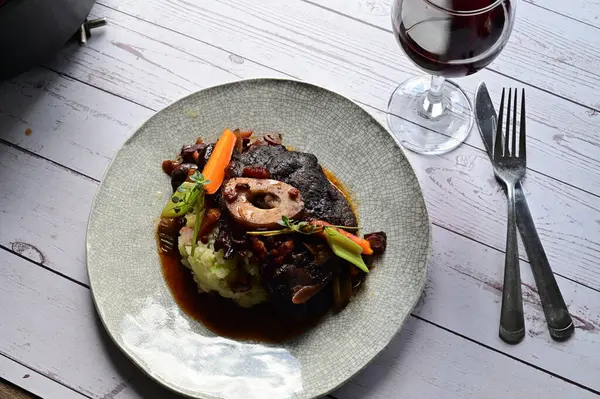 Sebzelerle servis edilen ve taze otlarla süslenen sığır eti osso buco 'sunun yakın çekimi. Kemik iliğiyle pişirilmiş yumuşak et, bu geleneksel İtalyan yemeğinin dokusunu ve derinliğini vurguluyor. 