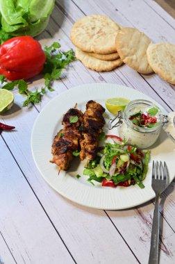Chicken kebab served with raita and fresh salad on a white plate, light background, healthy and delicious popular meal.
