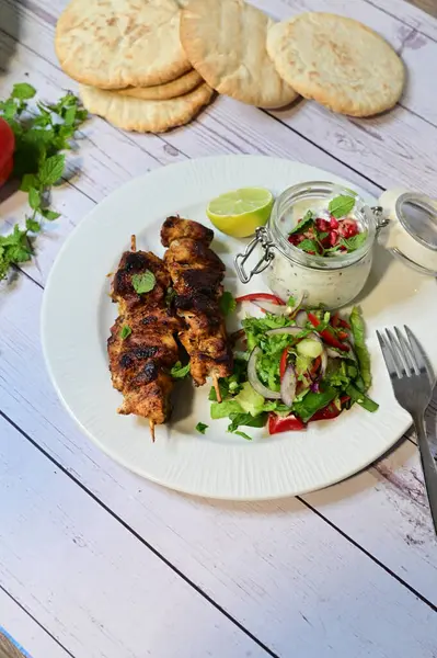 Chicken kebab served with raita and fresh salad on a white plate, light background, healthy and delicious popular meal.