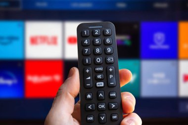 Bir adam arka planda bir televizyon ekranı olan akıllı bir televizyonun uzaktan kumandasını tutuyor ve bulanık bir video servis uygulama simgesi kullanıyor