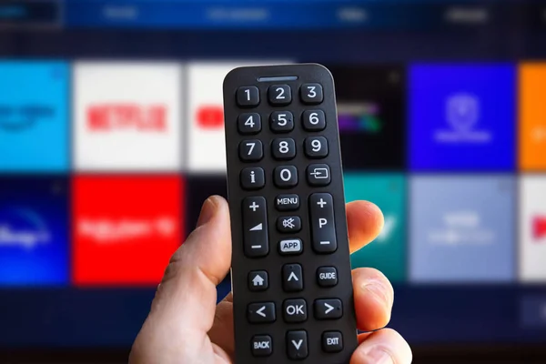 A man is holding the remote control of a smart TV with a television screen in the background with some blurry video streaming service app icon