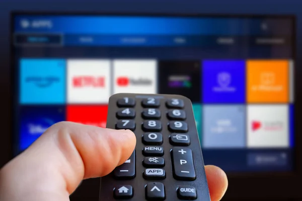 Bir adam arka planda bir televizyon ekranı olan akıllı bir televizyonun uzaktan kumandasını tutuyor ve bulanık bir video servis uygulama simgesi kullanıyor