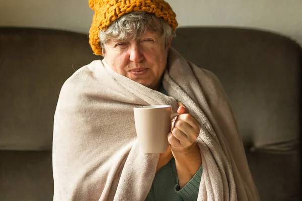 Örgü ören, battaniyeye sarılı ve sıcak çay içen yaşlı bir kadın. Yaşlı büyükannem evde üşüyormuş, evde ısıtma sorunu varmış. Avrupa 'da yoksulluk ve enerji krizi. Soğuk belirtileri.