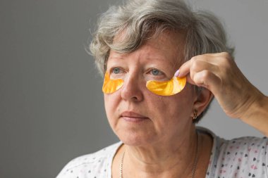 Gri arka planda izole edilmiş kırışıklıklara hidrojel göz bandı yapıştıran güzel bir kadının portresi. Kozmetik kullanan gri saçlı kadın, günlük sabah rutini. Yaşlanma ve cilt bakımı