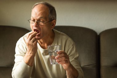 Beyaz kazaklı, beyaz giysili beyaz adam ağzına kırmızı ve sarı kapsül alıp camdan su içiyor. Yaşlı adam kendini hasta hissediyor ve evde ilaç alıyor. Sağlık ve terapi.