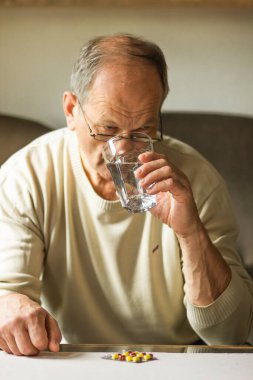 Beyaz kazaklı beyaz bir adam kapsülü alıp evdeki kanepede cam bardaktan temiz su içiyor ve hasta gibi hissediyor. Gözlüklü yaşlı adam ilaç alıyor. Sağlık ve terapi kavramı.