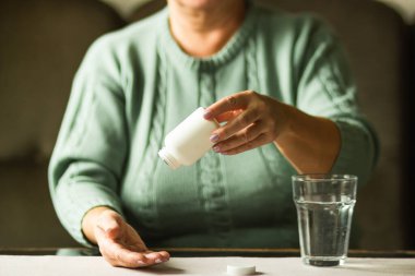 Yaşlı kadınların elleri beyaz plastik şişeyi haplarla tutuyor ve ilave hapları bir bardak suyla almaya hazır, ilaç alıyor. Hasta yaşlı kadın baş ağrısı ve soğuk algınlığına karşı ilaç istiyor..