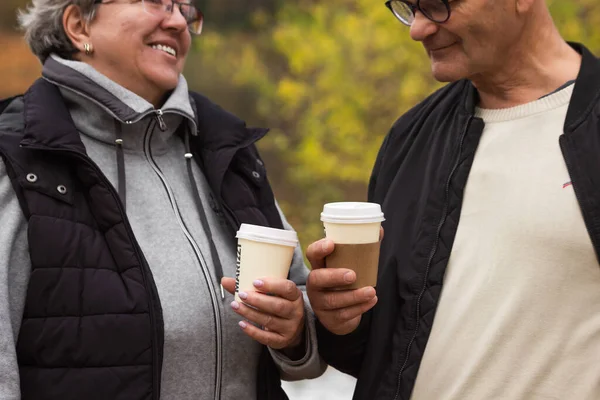 Happy senior couple walking in autumn park and drinking coffee. Smiling elderly couple talking and holding disposable cups, coffee to go. Weekend leisure activity for old people concept. Active life.