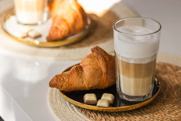 stock image Glass cup of cappuccino coffee or latte macchiato with french croissant and chocolate on saucer on the wicker lining. White table served for breakfast. Daily morning routine. Slow living concept.