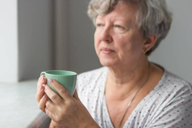 Beyaz gecelikli yaşlı kadın elinde naneli çay ya da kahve tutuyor, günlük sabah rutini. Gri saçlı bayan rahatlıyor ve sıcak içecek içiyor. Yaşlılar için boş zaman etkinliği konsepti.