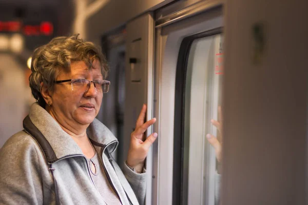 stock image Senior woman in glasses and gray sweatshirt traveling by train and looks out of the window. Portrait of happy old woman enjoying a train trip. Aging population and travel concept. Elderly tourist.