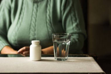 Masanın üzerinde haplar ve bir bardak su olan beyaz plastik bir şişe. Kıdemli kadın takviye hapı almaya hazır. Büyükannem ilaç alıyor. Hasta yaşlı kadın baş ağrısı ve soğuk algınlığına karşı ilaç istiyor..