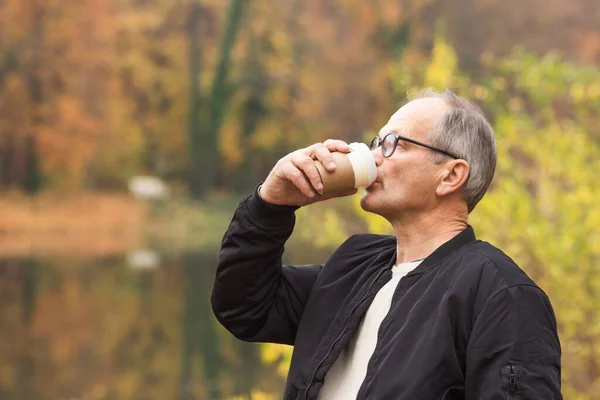 stock image Senior man in glasses and casual clothes walking and drinking hot coffee in autumn city park. Nature background. Elderly man holding disposable paper cup in his hands. Active seniors concept. 