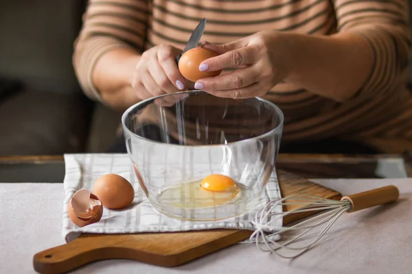 Old woman\'s hands cracking eggs into glass bowl with knife. Prepare for making omelet or pancakes for breakfast. Bowl of fresh eggs and whipper isolated on wooden cutting board and table background.