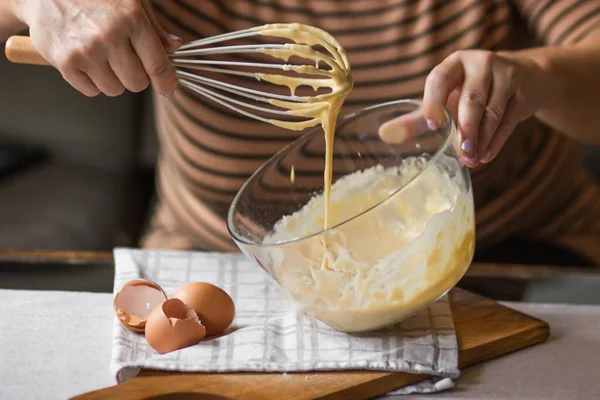 Senior woman beats eggs with flour for dough with a wisk in a glass bowl. Ingredients for a christmas pie or morning pancakes on kitchen table. Bowl of fresh batter. Cooking in process concept.