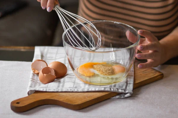 Elderly woman beats eggs with a wisk in a glass bowl. Prepare for making christmas pie or morning pancakes on kitchen table. Bowl of fresh eggs and whipper. Cooking in process concept.