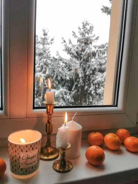 Burning candles in golden vintage candlesticks with group of mandarines at the window sill. Cozy winter day at home. Merry season. Window view on the snowy trees.