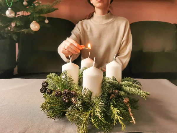 Woman in beige sweater lights candles on christmas advent wreath with matches. Handmade wreath with fir branches, tree cones and four candles. Horizontal photo.
