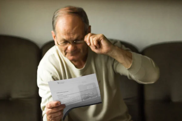 Worried senior man in white sweater and glasses reading utility bill in period of energy crisis. Old man checking domestic finances and stressed of debt. Financial problems and high heating prices.