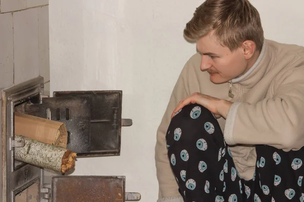 可怜的成年人穿着暖和的休闲装 在家里的旧火炉里烧柴烧纸 白人在寒冷的冬日里给他的房子取暖 贫穷和能源危机概念 — 图库照片