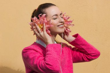 Pembe kazaklı gülümseyen esmer kadın çiçekli kulaklıklar takıyor ve müzik dinliyor. Pastel sarısı arka planda izole edilmiş. Dahlia Karpuz çiçek tomurcukları kulaklık niyetine. Yaratıcı fikir kavramı.