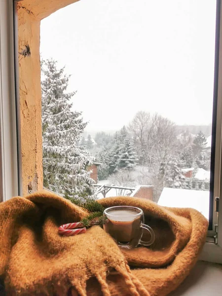 Window view to the snow covered trees. Ginger wool scarf and glass cup of hot coffee on the sill. Cozy winter morning at the cottage.
