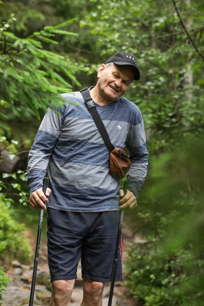 stock image Smiling senior man with trekking sticks for Nordic walking hiking on top of the mountain. Green nature background. Healthy lifestyle and active seniors concept. Summer journey.