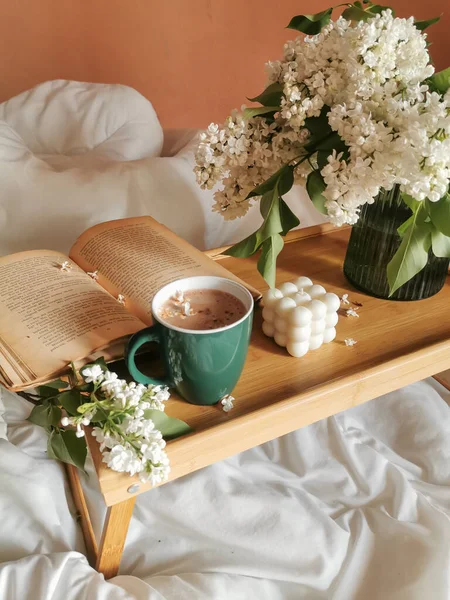 Spring still-life with folding wooden table in bed, green cup of coffe, open book, bubble cube soy candle and bunch of white lilac. Cozy morning at home. Remote education or quarantine concept.