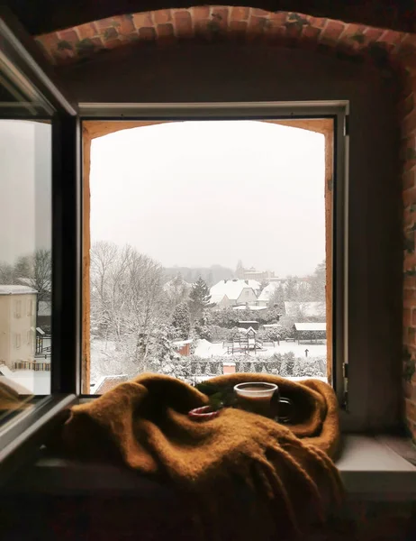 Window view to the snow covered trees and roofs. Ginger wool scarf and glass cup of hot coffee on the sill. Cozy winter morning at the cottage.