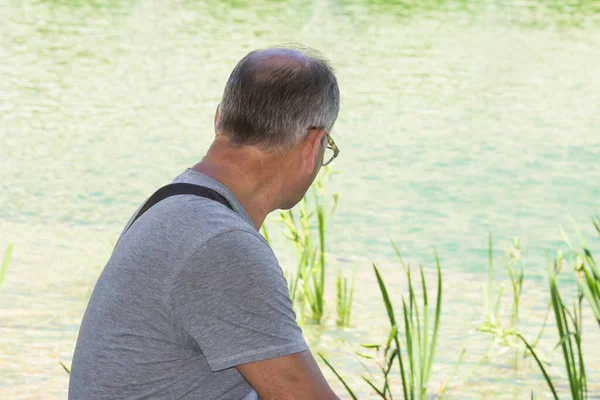 Çek Cumhuriyeti Prag şehir parkındaki çeşmenin yanında oturan gözlüklü ve gri tişörtlü son sınıf öğrencisi. Dikiz aynası. Emekli turist dinleniyor ve yeşil gölet suyuna bakıyor. Yaz yolculuğu konsepti.
