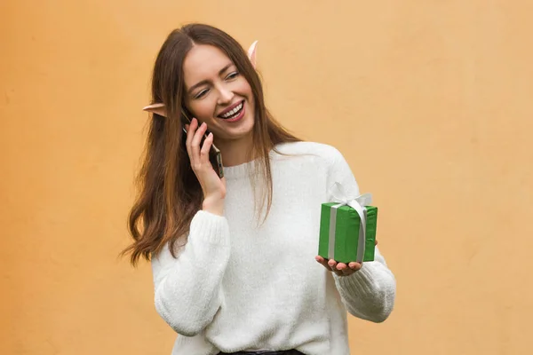 Smiling young woman with elf ears talking on mobile phone and holding green gift box in hand, against yellow wall background. New Year or Christmas online shopping and delivery concept. Communication.