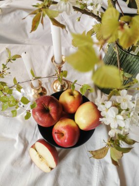 Bir kasedeki kırmızı ve sarı elmaların üst görüntüsü, yeşil yapraklı ve çiçekli bahar dalları, beyaz mumlarla süslenmiş klasik şamdanlar. Beyaz arka plan. Konforlu ev.