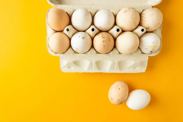 Top view of open egg box with ten farm eggs and two eggs near isolated on yellow background. Fresh organic chicken eggs in carton pack. Easter holiday concept.
