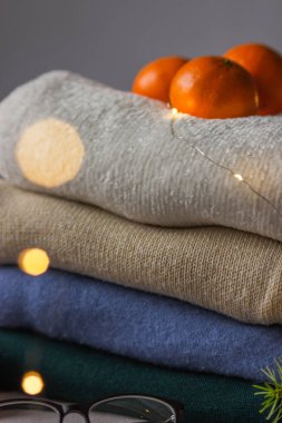 Farklı renklerde katlanmış kazaklar köknar dalı, mandalina ve gözlüklerle mükemmel bir şekilde üst üste istiflenmiş. Bokeh ışığı. Kış ya da sonbahar sıcacık kadın kıyafetleri. Tatil günleri sihirli bilgelik.