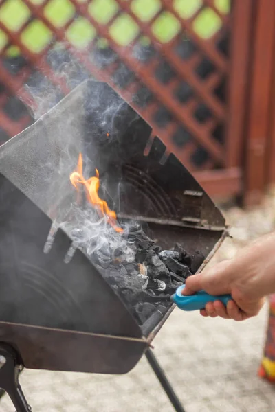 Male hand kindles fire in grill with long blue gas lighter. Man holding and using BBQ lighter to start fire for charcoal in folding portable brazier. Smoke and flame. Barbeque time on backyard concept