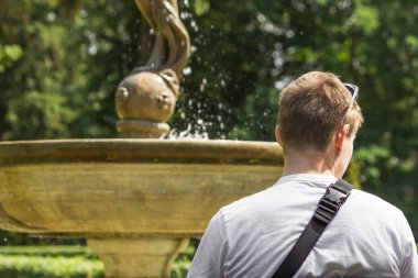 Genç sarışın, yalnız adam yaz parkındaki çeşmenin önünde bankta oturuyor. Arkadan bak. Beyaz tişörtlü turist doğada tek başına vakit geçirip hayatı düşünüyor. Ruh Sağlığı.