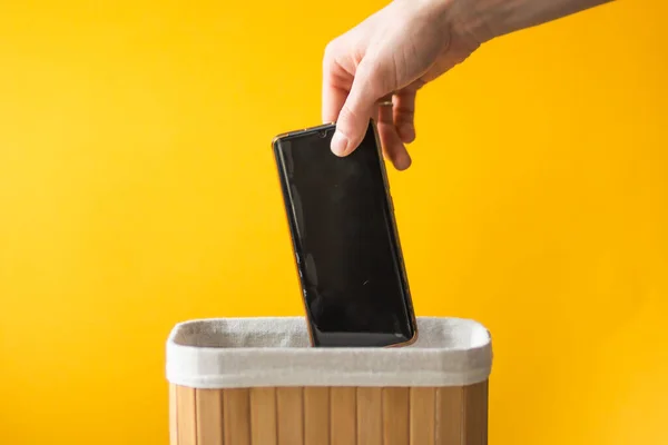 Man\'s hand putting smartphone into wooden basket on a yellow background. Dependance and digital detox concept. Social media addiction. Throw out your gadget in a trash can, free from mobile phone.